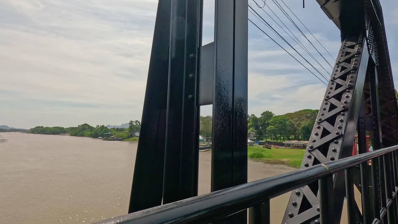 Point-of-view footage moving across a steel railway bridge over the River Kwai in Kanchanaburi, Thailand, under bright daylight with steady camera movement