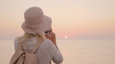 young woman tourist in a pink hat makes a photo of the rising sun on the sea