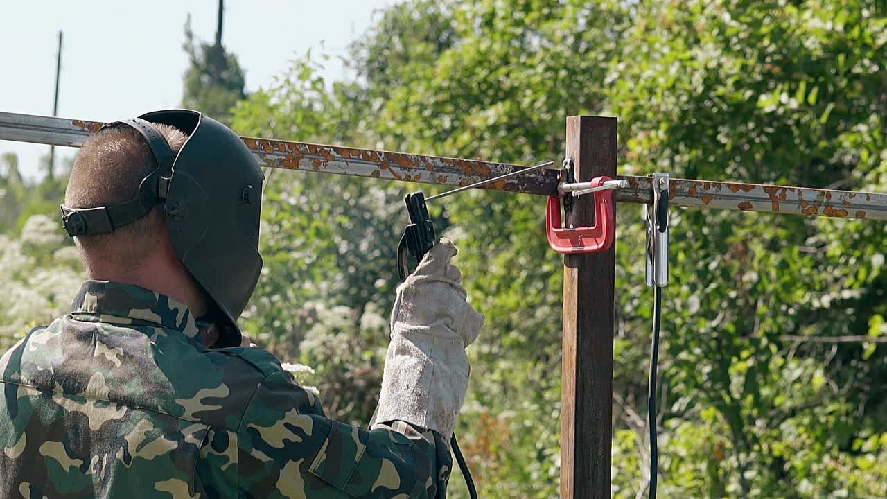 천천히 움직이는 뒷면 뷰 빌더는 녹색 마당에서 울타리를 용접합니다.