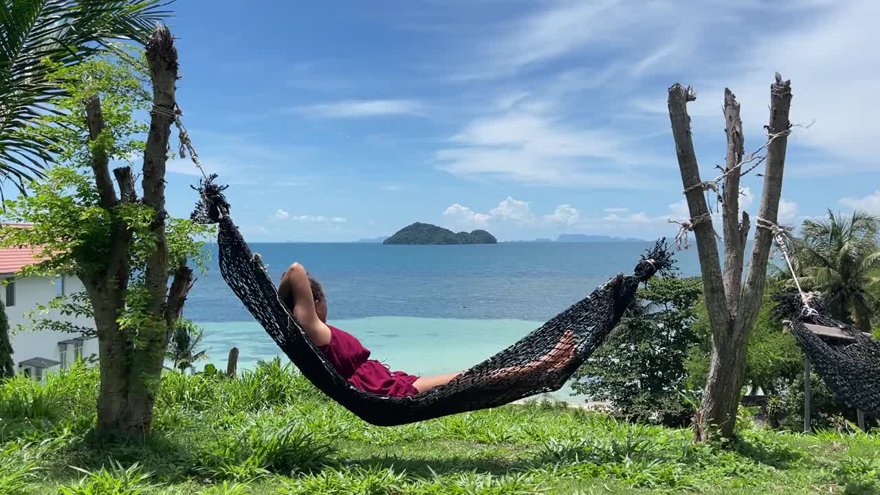 A person relaxes in a black hammock, enjoying the serene view of the turquoise ocean and distant islands, near Nui Beach in the Phi Phi Islands, Thailand.