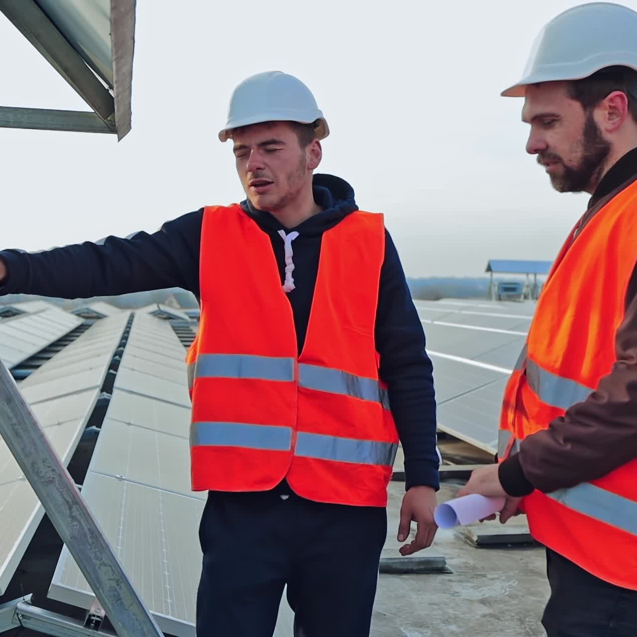 Workers near the modern electricity equipment. Technicians in orange uniform talking about the installation on the solar panels background.