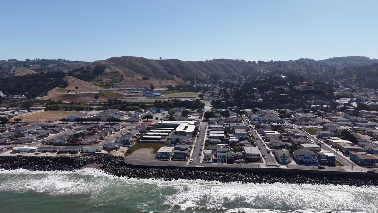 Luftfoto af kystbyen Pacifica, Californien, viser huse langs Stillehavskysten med bølgende bakker i baggrunden på en lys, solrig dag med klar blå himmel