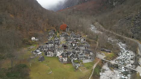 Drone-strafing-above-the-village-in-Cavergno-showing-the-stone-houses,-a-bridge-above-Maggia-River,-and-the-misty-mountains-in-Vallemaggia-district,-in-the-canton-of-Ticino-in-Switzerland