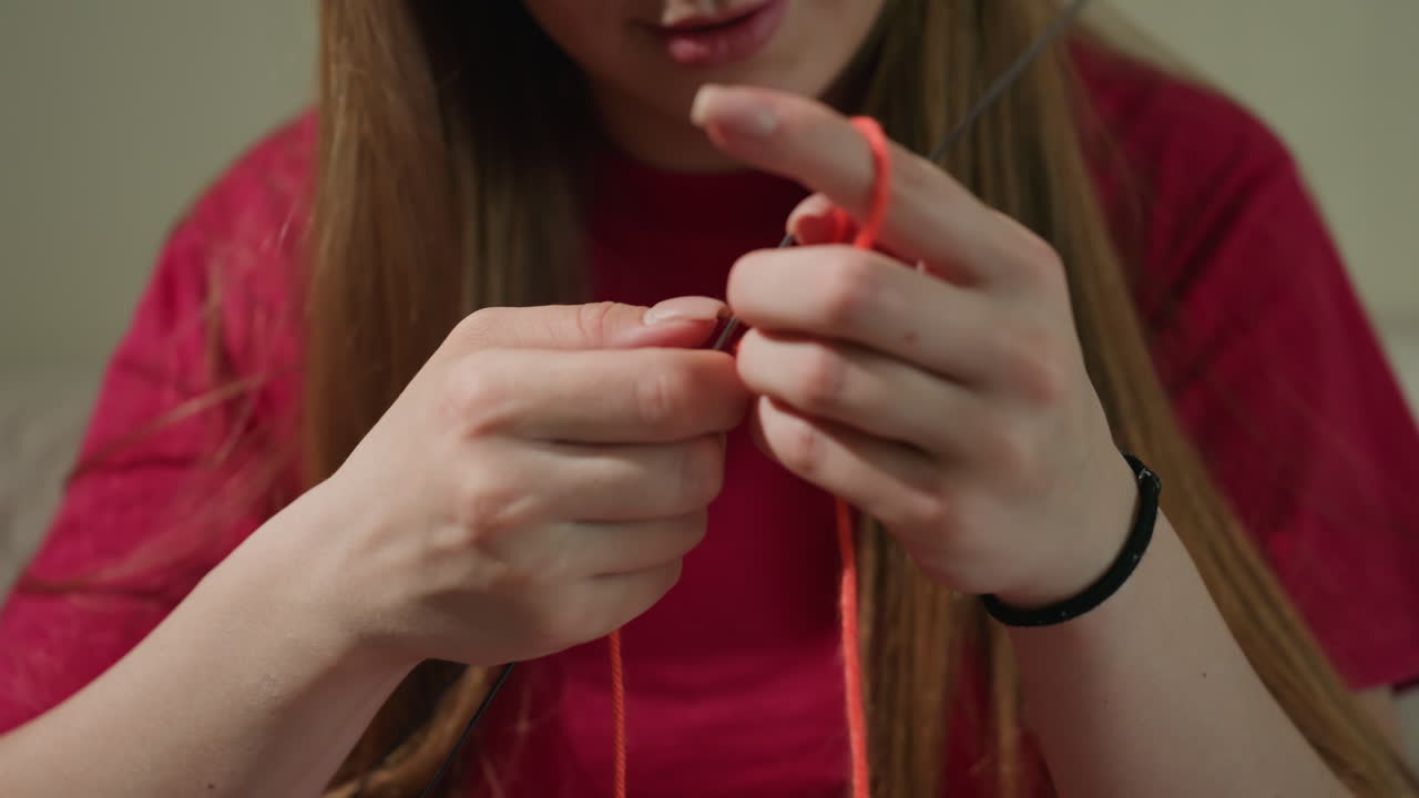 젊은 여성이 털실로 작품을 만들고 있어요. 한 여성이 여가 시간에 실내에서 오렌지색 털실로 평화롭게 뜨개질을 하고 있으며, 젊은 여성은 아늑하고 평온한 분위기 속에서 편안하게 뜨개질을 즐기고 있어요.