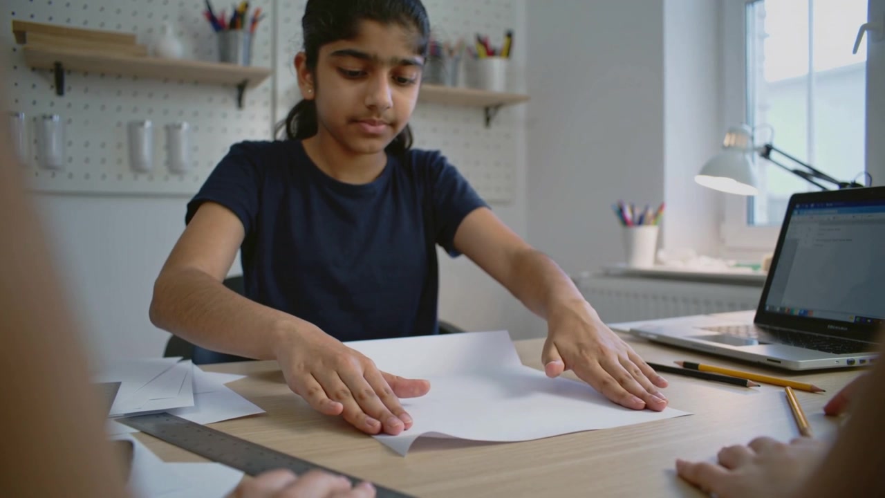 Smoothing teen navy tee aligning folding paper at home desk, second hands entering to help craft