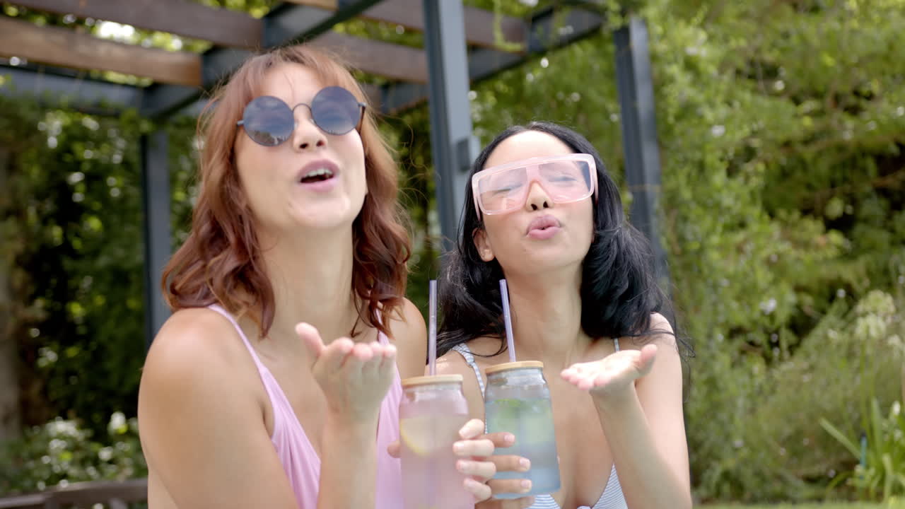 Two biracial female friends blowing kisses by poolside at home, holding drinks