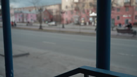 a close bokeh view of a city street, focusing on a blue railing with a blurred view of people walking across the road in black clothing and cars passing by