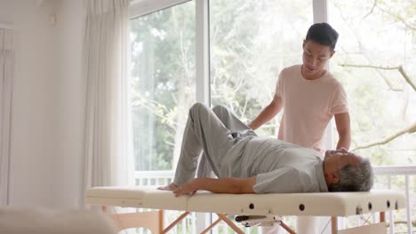 diverse male physiotherapist advising and senior male patient exercising on couch, in slow motion