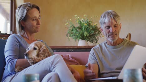 happy senior caucasian couple sitting in living room at home with pet dog using tablet and talking