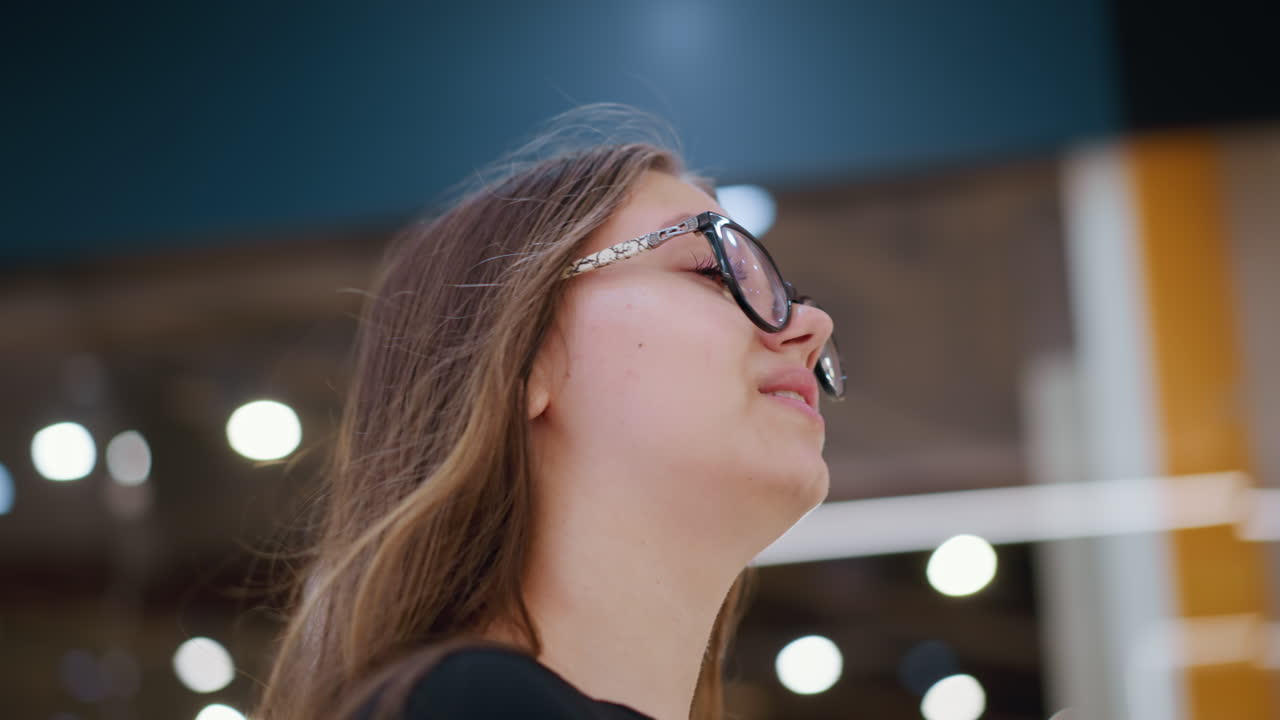 primer plano de una mujer con gafas sonriendo ligeramente mientras mira hacia arriba con luces bokeh suaves en el fondo, la inscripción borrosa en la pared mejora la atmósfera moderna de venta al por menor con una iluminación cálida