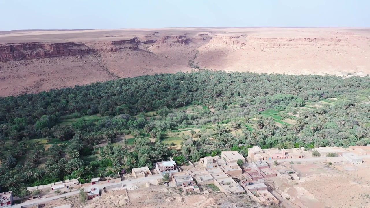 Vista aerea con drone di un villaggio tradizionale che si affaccia sull'Oasi di Tafilalt, dove vasti palmeti di datteri e campi fertili prosperano sotto i suggestivi altipiani desertici del Marocco sud-orientale