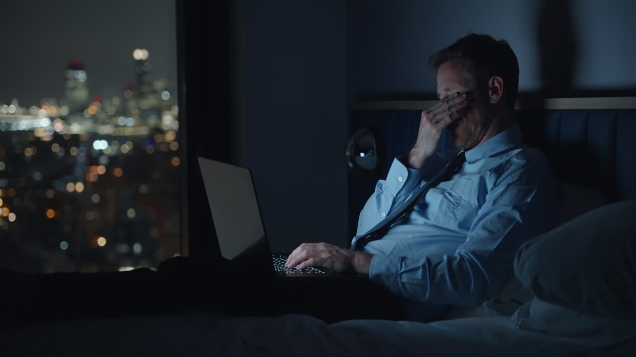 Tired businessman typing on his laptop while sitting in bed in hotel or apartment at night overlooking the city