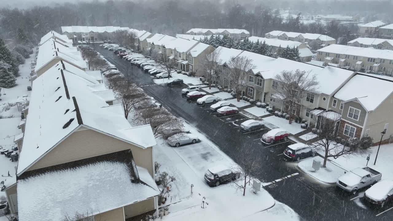 Vista aérea de un complejo residencial suburbano durante una fuerte tormenta de nieve invernal, con casas adosadas cubiertas de nieve, coches aparcados y nieve cayendo. Fría atmósfera estacional en un pequeño asentamiento de EE. UU. Toma descendente