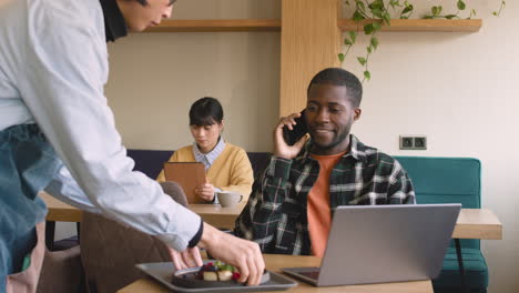 homme assis à table dans un café et parlant au téléphone portable pendant que le serveur lui sert de la nourriture puis prend la commande d'un autre client