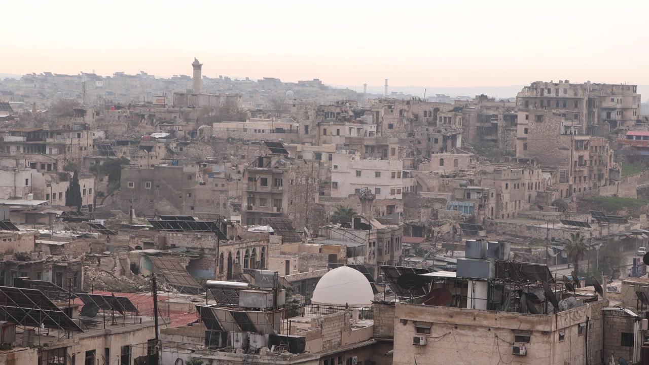 Hazy panoramic view of the Aleppo skyline in Syria, featuring traditional minarets and urban rooftops. A vast landscape of the ancient city under a dusty morning sky