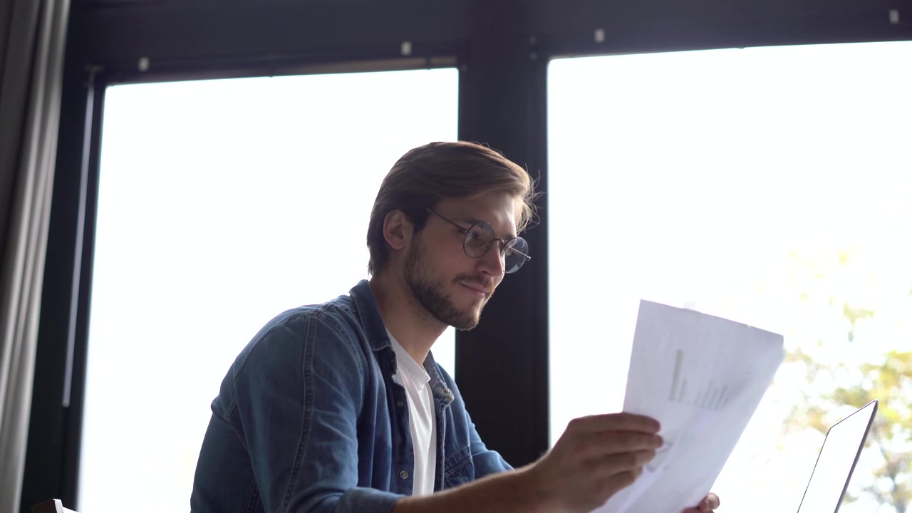Young Man Working on Laptop and Reviewing Documents in Modern Office