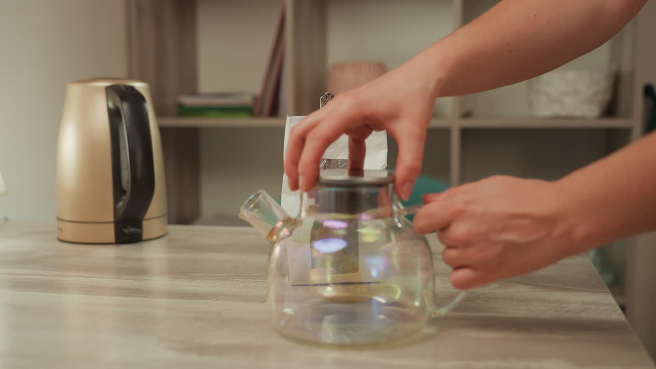 Iridescent glass teapot on countertop, esthetician hands placing filter and lid, electric kettle in background, gentle steam ready, serene spa lighting, preparation for herbal infusion