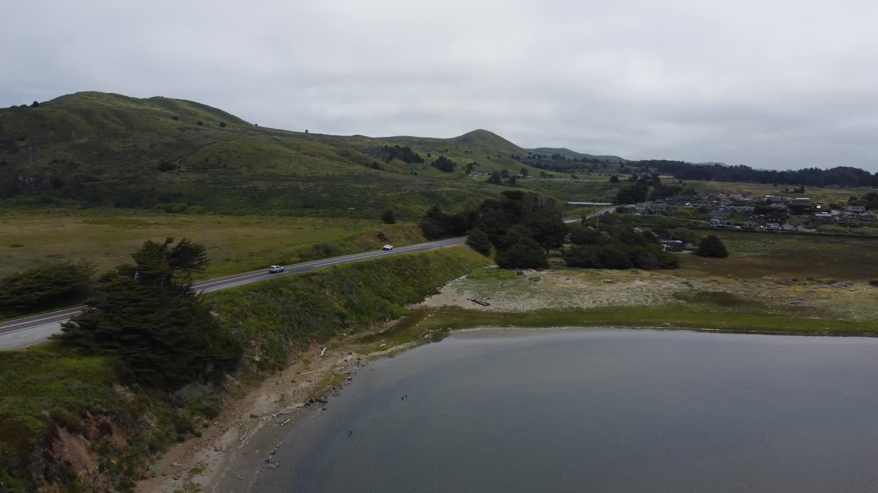 Cars driving down the road on a road trip near the coast of Northern California. Road is surrounded by green hills, trees. and water from the nearby ocean. Filmed on an overcast day with 4K drone.