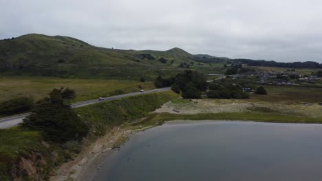 cars driving down the road on a road trip near the coast of northern california. road is surrounded by green hills, trees. and water from the nearby ocean. filmed on an overcast day with 4k drone.