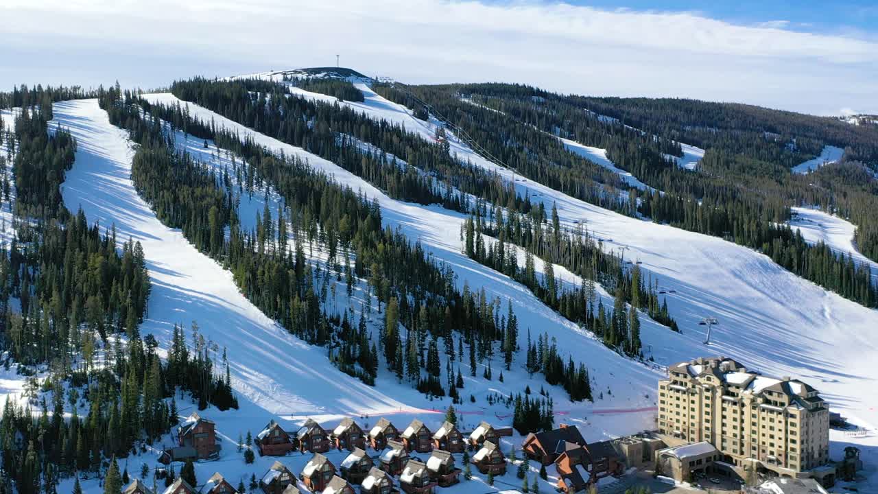 Winter drone shot above Big Sky, Montana reveals ski resort views, snow-draped trees, and cozy rental condos tucked into the mountainside, perfect for a scenic alpine vacation retreat