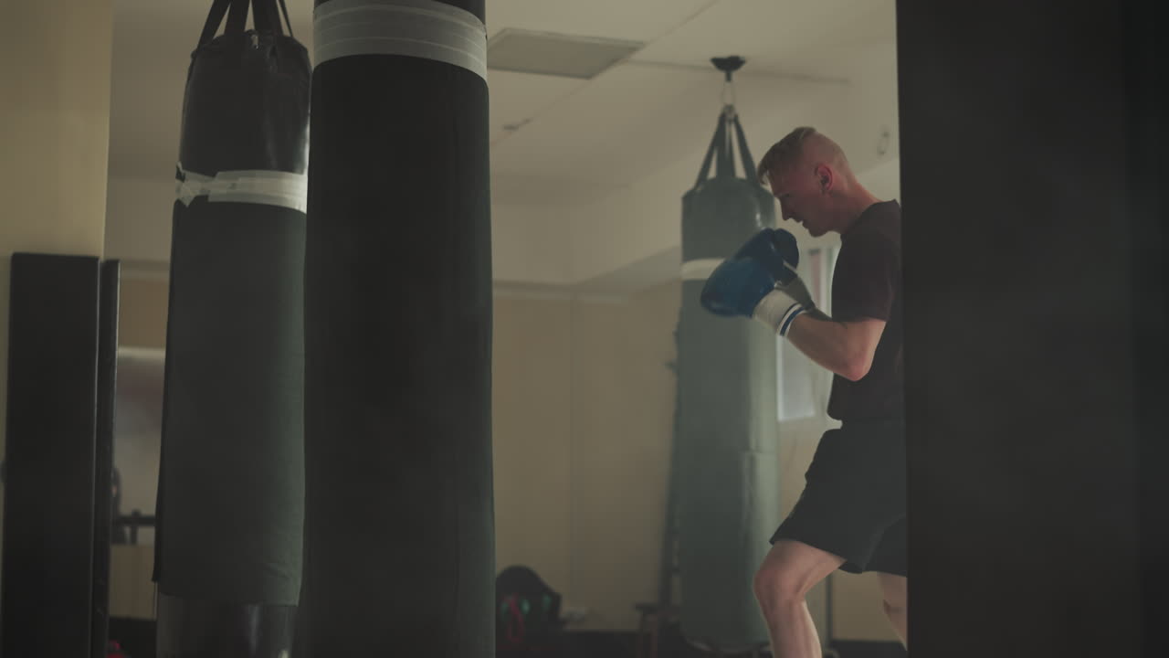 Athlete hits punching bag by fists and legs in gym. Muscular man exercises kick on equipment in fighting club slow motion. Martial arts trainer