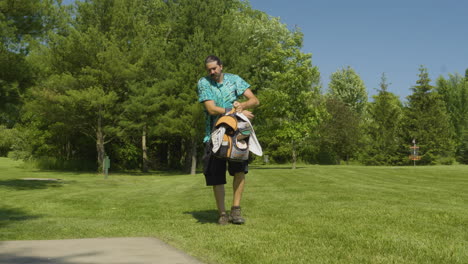 A-man-prepares-his-disc-golf-bag-on-a-sunny-day-in-a-lush-park,-surrounded-by-vibrant-greenery