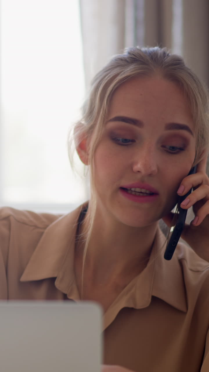 Young blonde woman in beige shirt talks on mobile phone drinking cup of tea near laptop at table against bright window slow motion