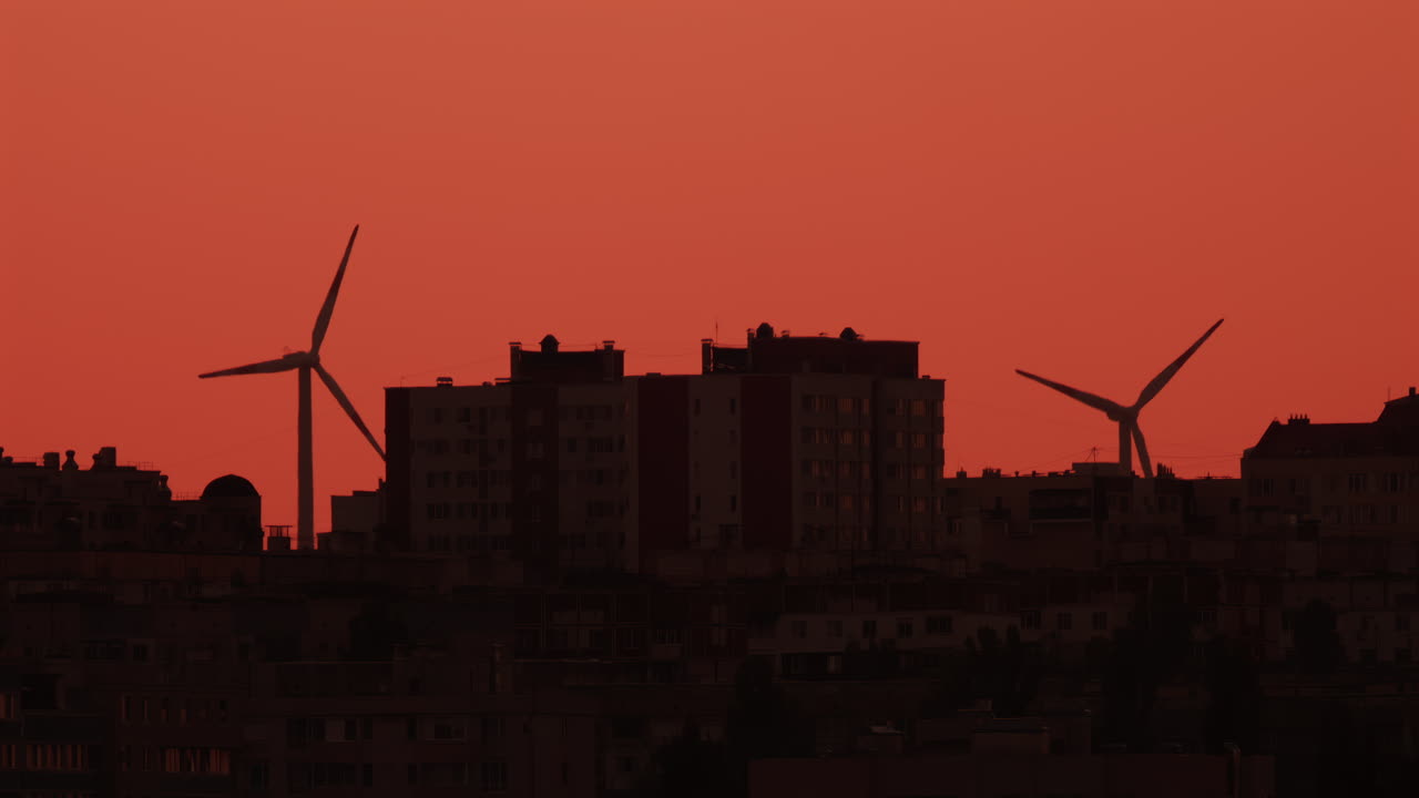 Two wind turbines stand tall near city buildings under an orange sky