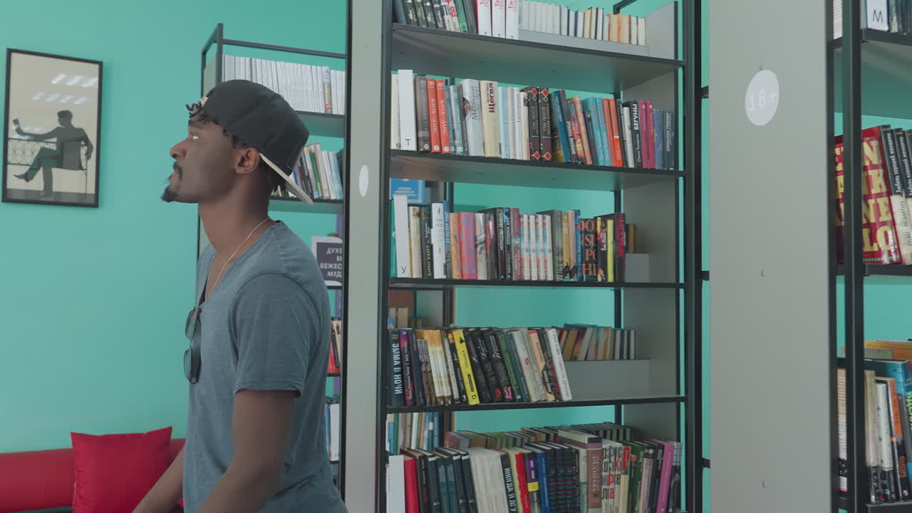 Young man wearing cap and casual shirt browsing bookshelves in library, focusing on selection with interest while standing between rows of colorful books in quiet study environment