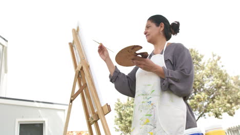 Feliz-Mujer-Mayor-Birracial-Pintando-Cuadros-Y-Sonriendo-En-Un-Jardín-Soleado,-Cámara-Lenta,-Espacio-De-Copia