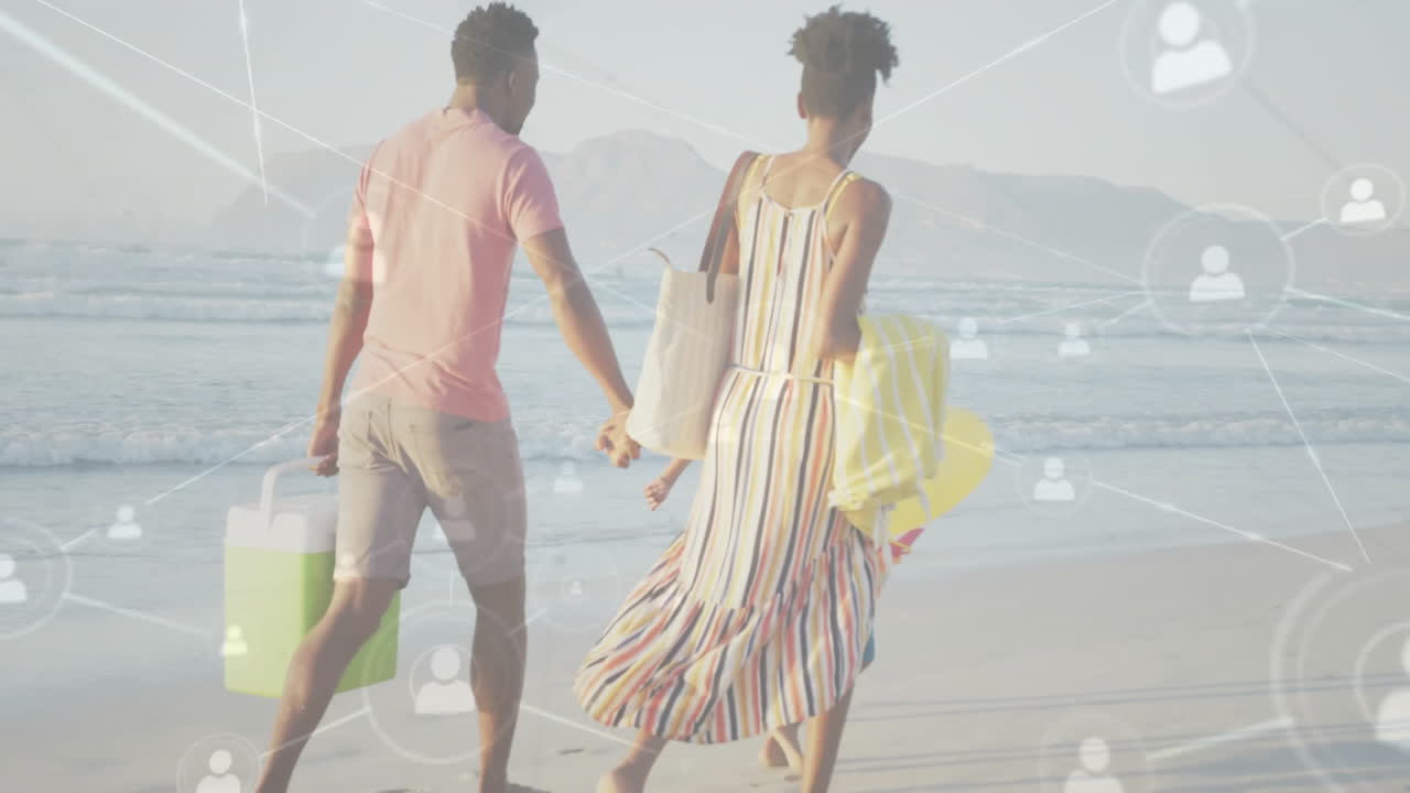 Couple walking on sandy beach, carrying cooler and float, displaying digital technology icons