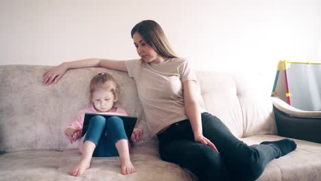 Young-mother-and-daughter-are-sitting-on-the-couch-and-training-using-a-tablet-computer