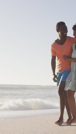 smiling african american couple embracing and walking on sunny beach