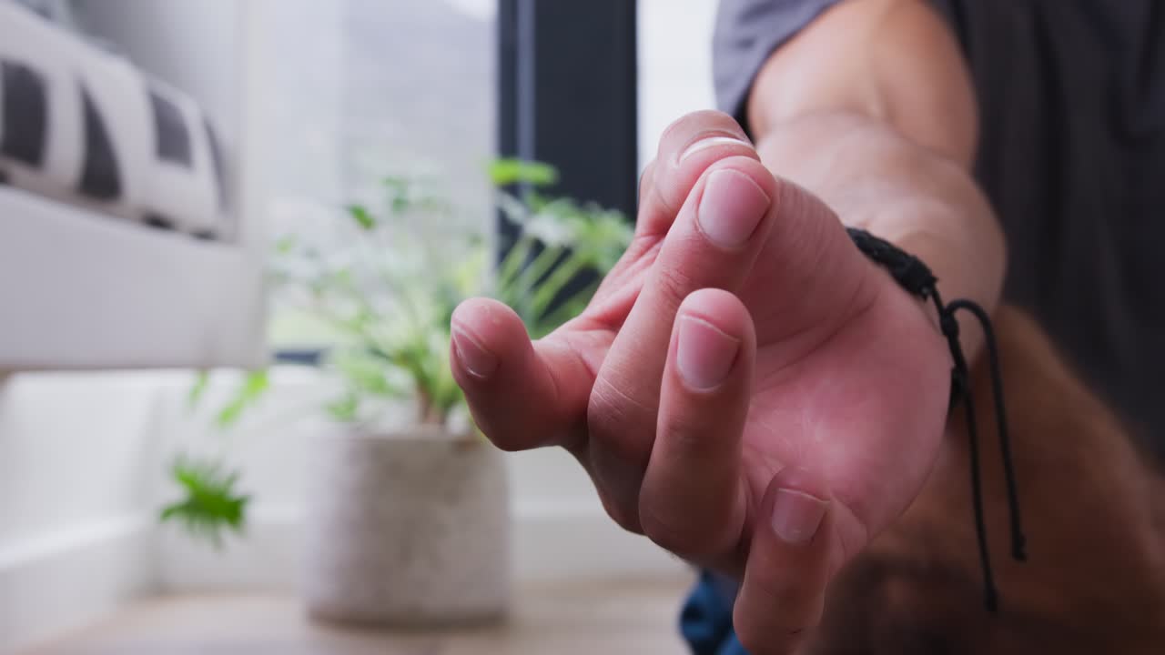 Lens focusing on hand, man rotating, pressing palms together by window, plant, couch in meditation