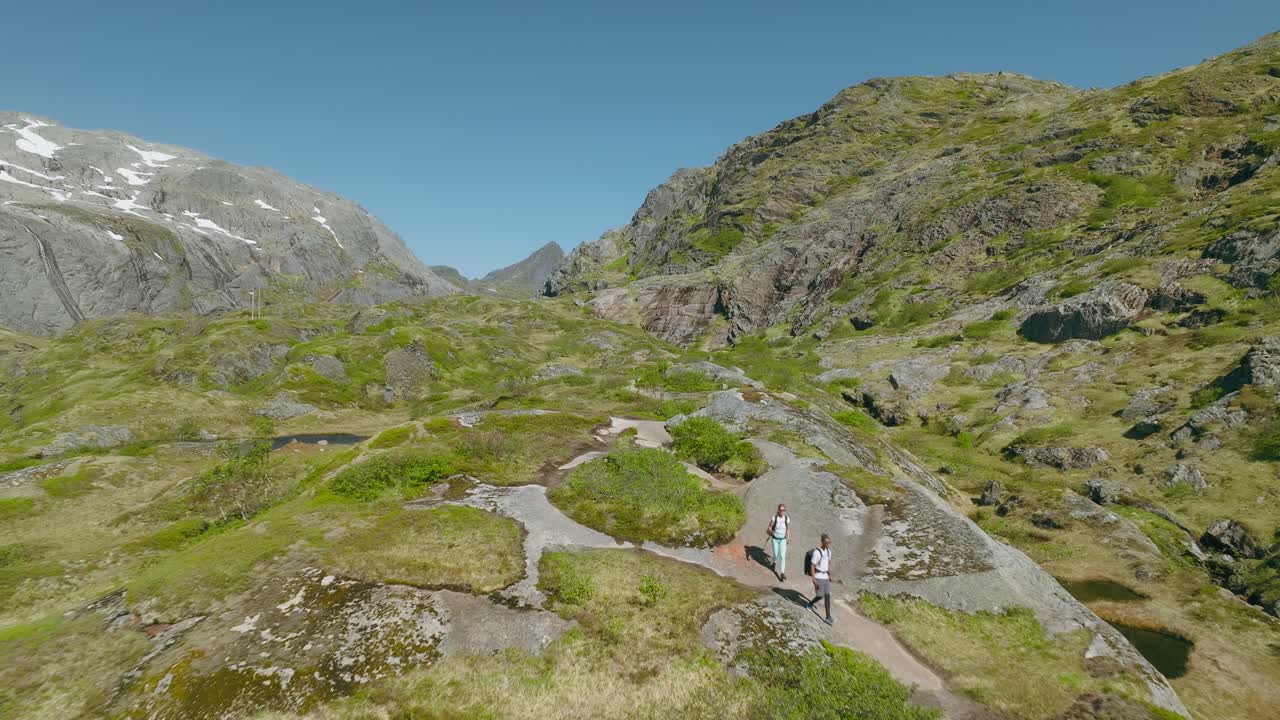Para wędruje przez surowy, skalisty krajobraz między granitowymi górami na Lofotach w Norwegii, pod czystym, błękitnym niebem, ukazując przygodę i malownicze piękno nordyckie.