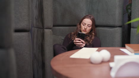 young woman dressed in black sits on cushioned booth with legs crossed while operating phone, wooden table with decorative items placed beside her, relaxed indoor setting with calm focused expression