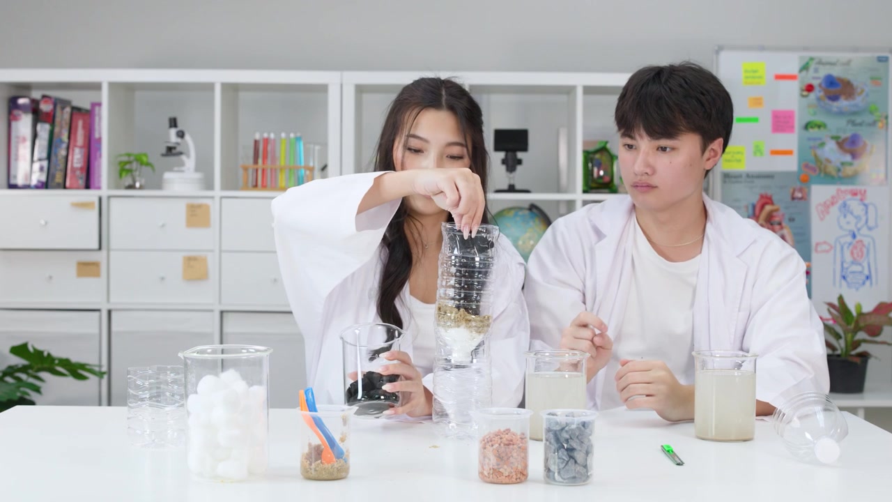 นักเรียนสองคนในชุดเสื้อกาวน์กำลังประกอบเครื่องกรองน้ำ DIY โดยใช้ขวดพลาสติกและถ่าน