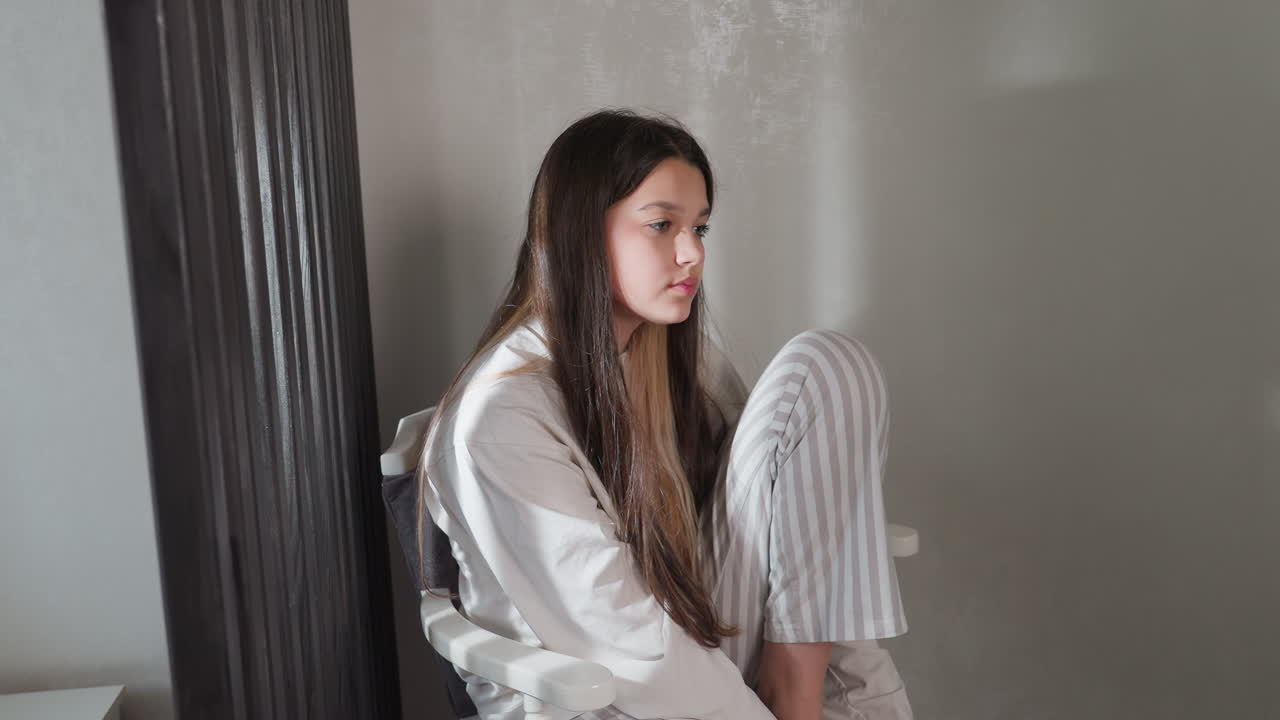 Female student sits quietly in striped pajamas on chair, knees hugged, long brunette hair falling, thoughtful gaze toward blank wall, soft indoor light, wood room divider beside