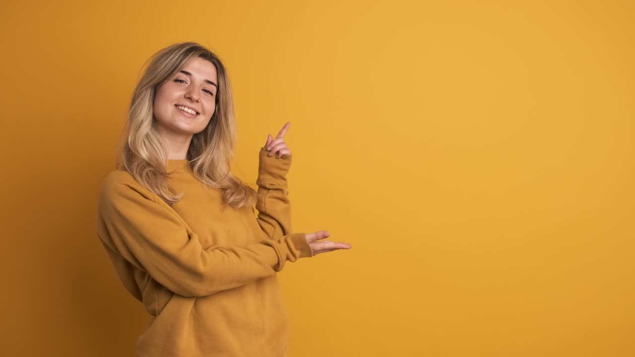 Cheerful young woman showing side space with fingers on yellow background