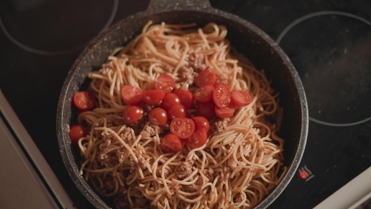 Primo piano dall'alto di spaghetti cotti conditi con pomodorini ciliegino dimezzati raggruppati al centro di una padella sul fornello, salsa lucida che ricopre la pasta, luce calda della cucina, un pasto casalingo rustico pronto per