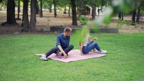 A-young-couple-goes-to-the-park-for-a-picnic.-The-guy-and-the-girl-spread-a-blanket-in-nature-and-sit-down-to-rest