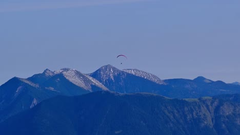 aerial-view-of-paraglider-with-red-parachute-flying-circes-on-a-bright-blue-sunny-day-in-front-of-snow-covered-mountain-peaks-in-alps