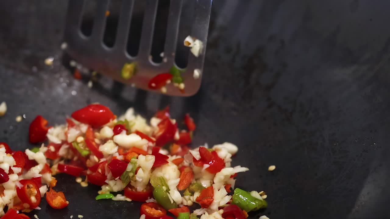 Close-up of a spatula stirring chopped chilli and garlic in a wok. Bright lighting highlights vibrant colors and dynamic cooking action