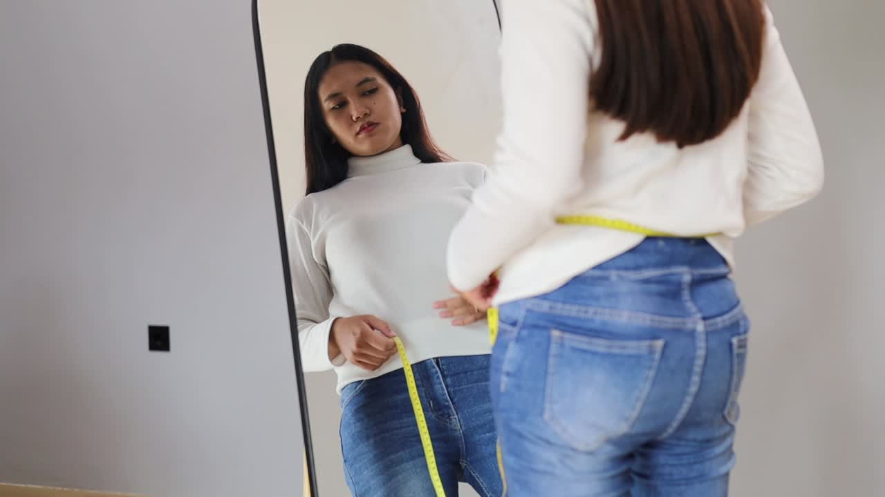 Unhappy Asian woman measuring waist near mirror, upset with visible weight gain