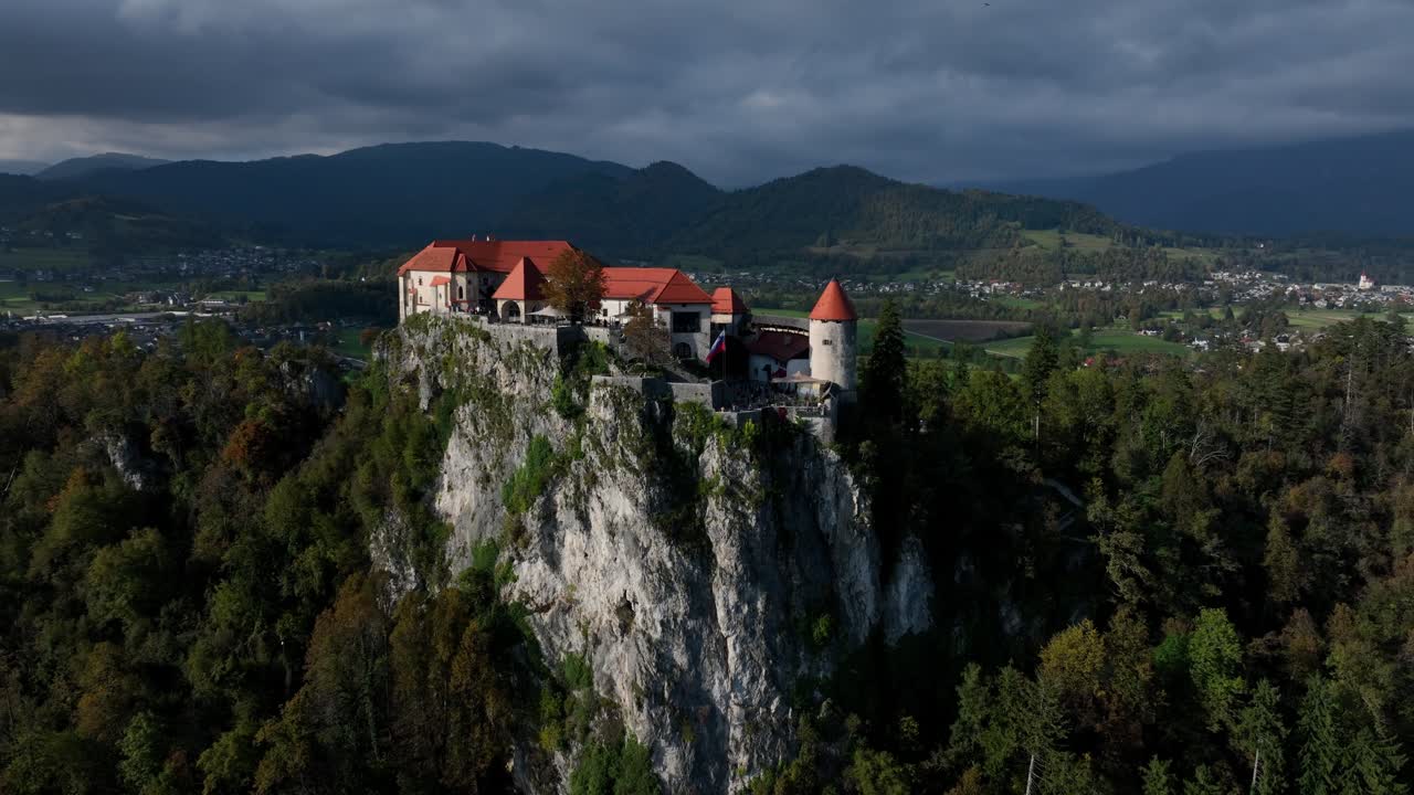 Gün doğumu sırasında kasvetli bulutların altında, vadiye ve dağlara bakan dramatik bir uçurumun tepesindeki Bled Kalesi, havadan orta yörüngeli çekim.