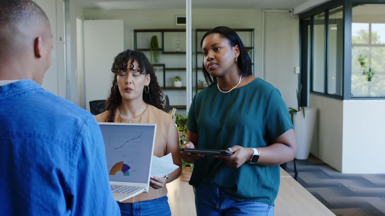Diverse female coworkers presenting laptop chart clarifying data at long table with man approaching