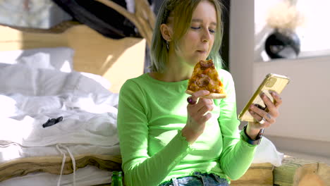 young blonde woman using mobile phone and eating pizza while sitting on the floor in the bedroom at home 1