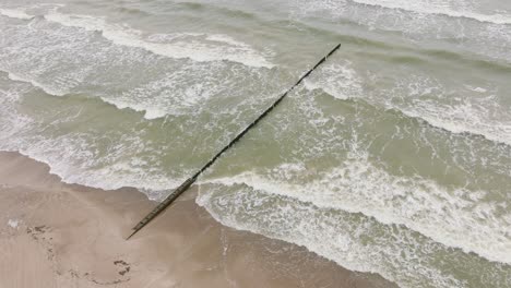 aerial birdseye view of baltic sea coast on a overcast day, old wooden pier, white sand beach, large storm waves crushing against the coast, climate changes, wide drone shot