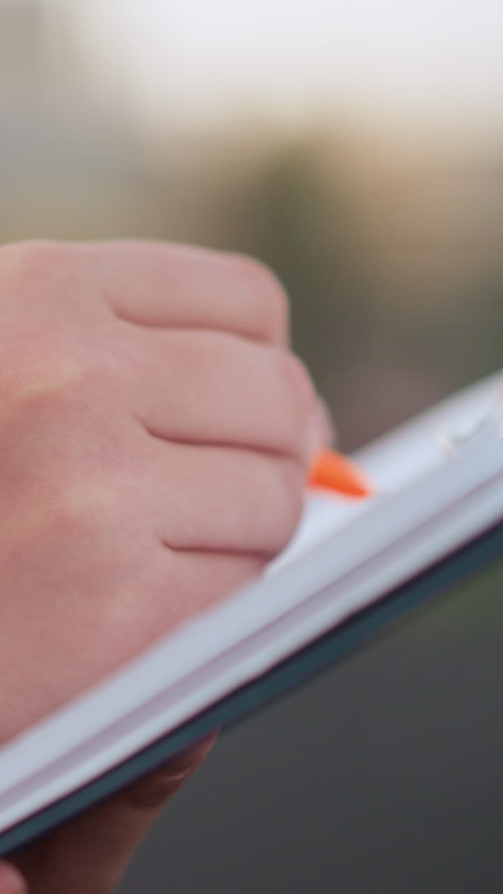 Primo piano laterale di una persona con maglia bordeaux che prende appunti su un blocco appunti all'aperto, usando una penna arancione; sullo sfondo si vede un campo sportivo con persone che camminano