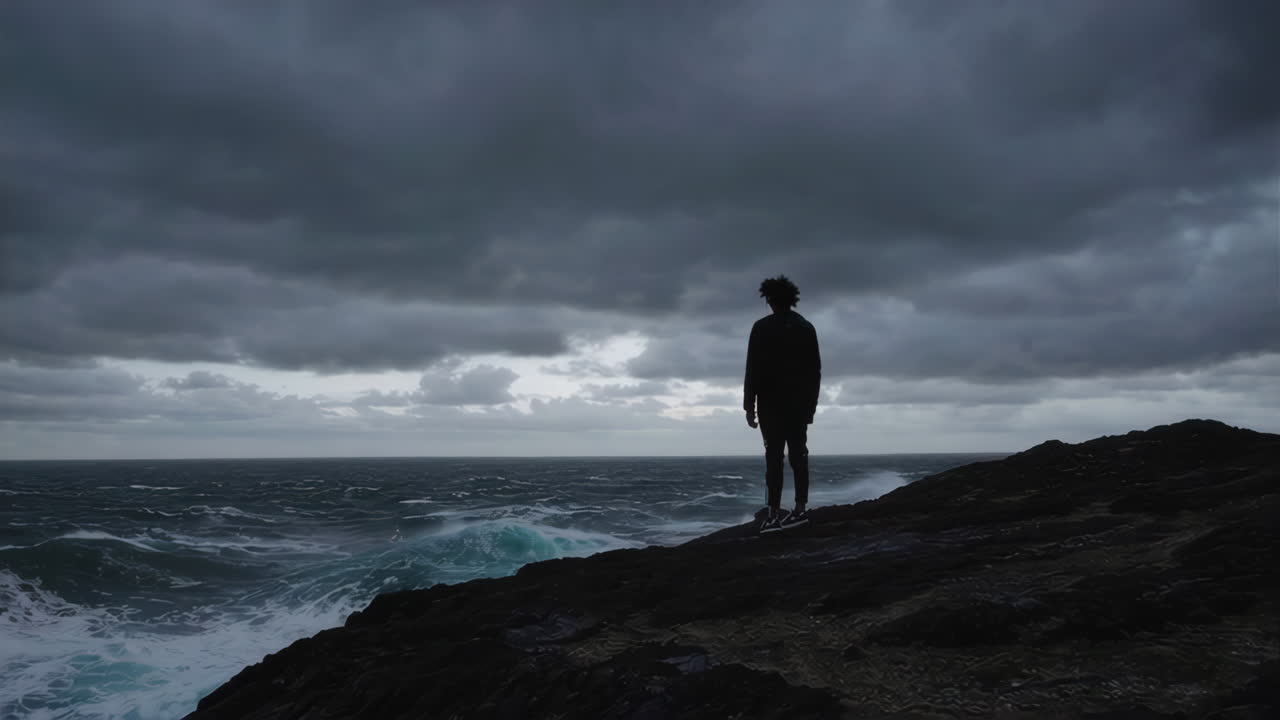 Person Standing on a Rocky Cliff Facing a Stormy Ocean